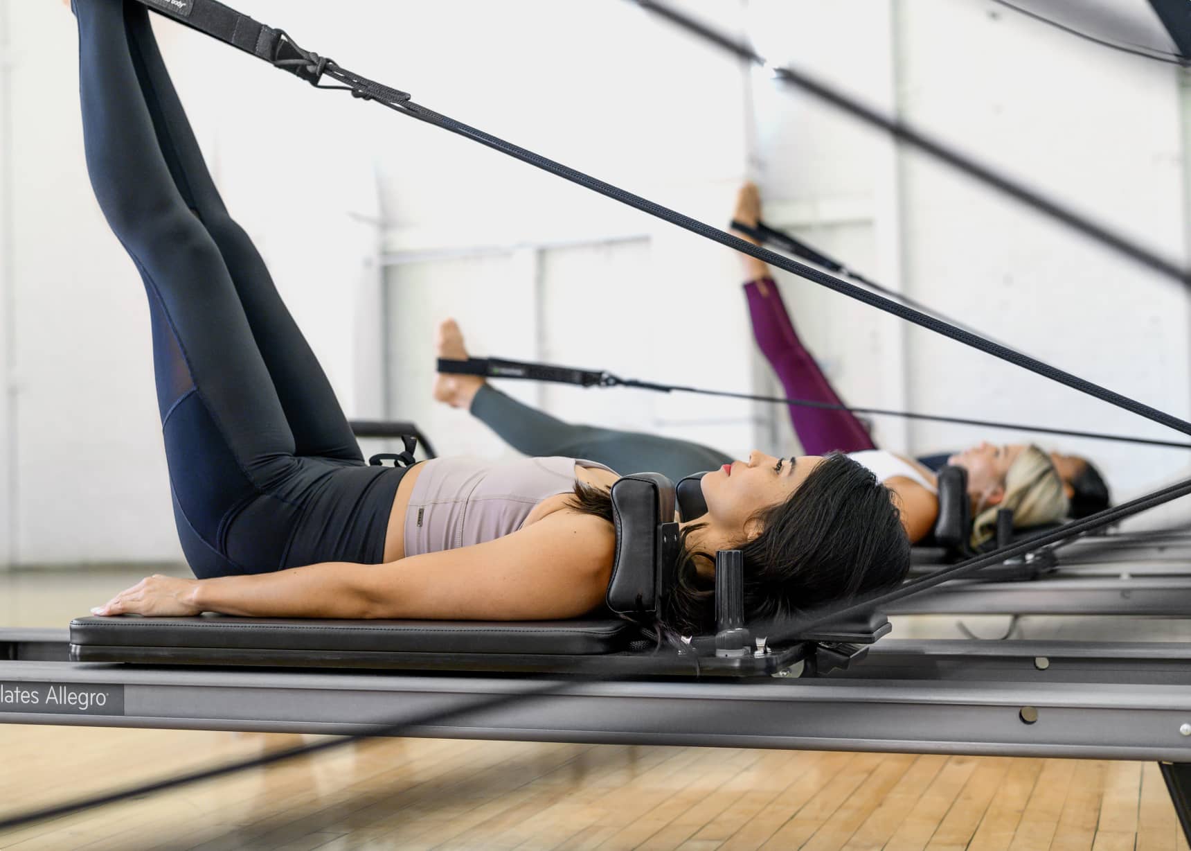 Side view of Pilates students in a class laying on reformers extending their legs with feet in straps